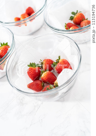 Bright red strawberries, interspersed with signs of mold, rest in a glass bowl lined with a paper towel on a white napkin, indicating improper storage techniques. 113194736