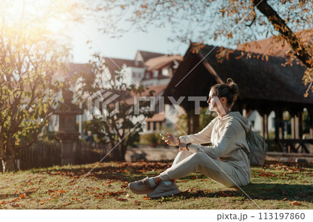 Young Fashionable Teenage Girl With Smartphone In Europian Park In Autumn Sitting At Smiling. Trendy Young Woman In Fall In Park Texting. Retouched, Vibrant Colors. Beautiful Blonde Teenage Girl 113197860