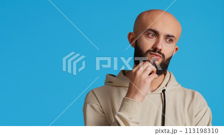 Thoughtful person being puzzled about a decision, standing over blue background in studio and thinking. Pensive middle eastern man being uncertain and doubtful about something. Camera 2. Thoughtful person being puzzled about a decision, standing over blue background in studio and thinking. Pensive middle eastern man being uncertain and doubtful about something. Camera 2. 113198310