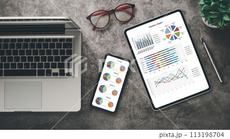 A sleek workspace showcasing a laptop, tablet with summary report, smartphone with charts, alongside eyeglasses and a green plant. 113198704