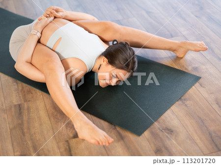 Happy young woman doing difficult pose of yoga, Tortoise Pose on mat in yoga studio 113201720