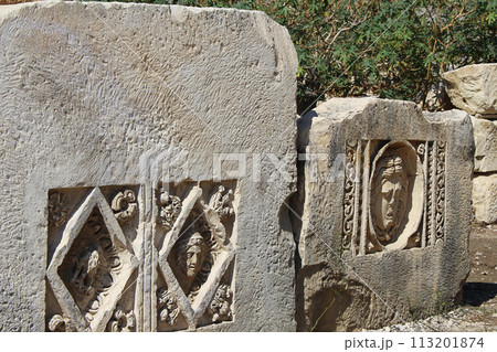 Historical stone bas-relief with carved faces in ancient city of Myra on sunny day outdoor. Dead civilization. Ruins of rock tombs in Lycia region, Demre, Antalya, Turkiye Historical stone bas-relief with carved faces in ancient city of Myra on sunny day outdoor. Dead civilization. Ruins of rock tombs in Lycia region, Demre, Antalya, Turkiye 113201874