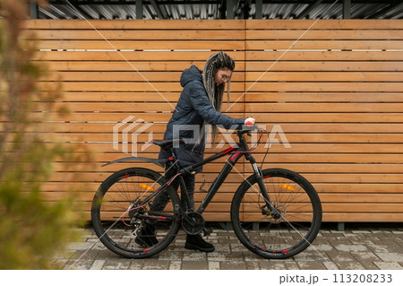 Cute young woman with dreadlocks hairstyle dressed in a winter jacket rides a bicycle 113208233