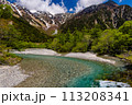 Clear, fast flowing mountain river in a forested valley in front of snowy peaks 113208341