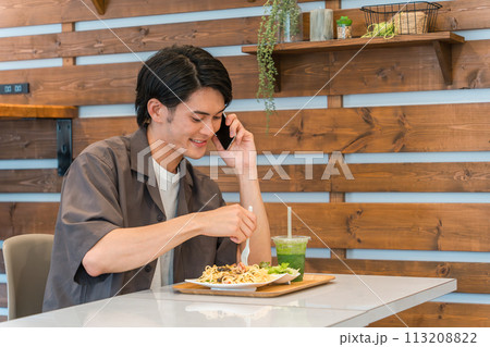 カフェで食事中にスマホで通話・電話する若いアジア人男性(ながらスマホ) カフェで食事中にスマホで通話・電話する若いアジア人男性(ながらスマホ) 113208822