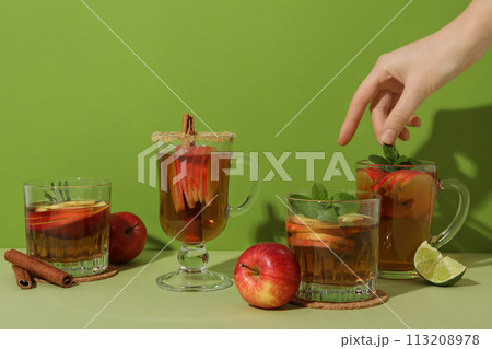 Glasses with apple cider, female hand, cinnamon sticks, piece of lime and red apples on green background 113208978