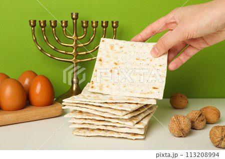 Matzo in hand, nuts, eggs on wooden stand and hanukkiah on green background Matzo in hand, nuts, eggs on wooden stand and hanukkiah on green background 113208994