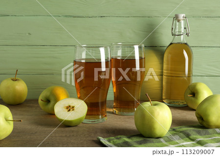 Glasses and bottle with apple cider and green apples on table on wooden green background Glasses and bottle with apple cider and green apples on table on wooden green background 113209007