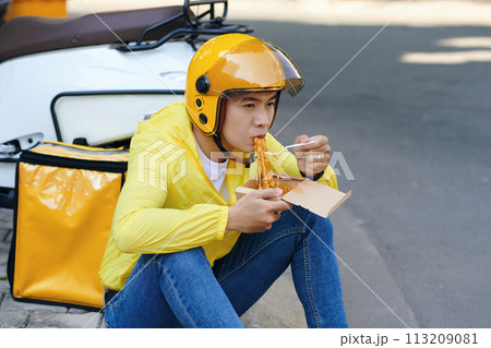 Pensive courier sitting on road side and eating noodles for lunch Pensive courier sitting on road side and eating noodles for lunch 113209081