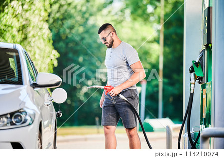Driver with gasoline pump refilling car gas tank. Confident man refueling his luxury white auto. Man in casual clothes and sunglasses at modern gas station. 113210074
