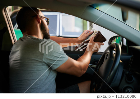 Young man paying for gasoline in cash. Man taking out money from wallet to pay for fuel. High fuel prices concept. Young man paying for gasoline in cash. Man taking out money from wallet to pay for fuel. High fuel prices concept. 113210080