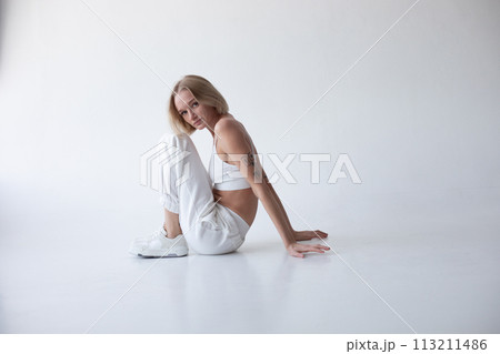 Beautiful blonde girl in a white top and tights posing on a white background. Sits on the floor. 113211486