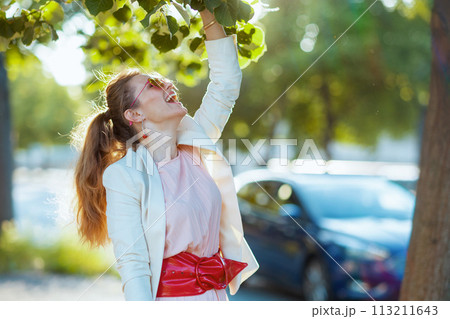 smiling trendy woman in pink dress and white jacket in city smiling trendy woman in pink dress and white jacket in city 113211643