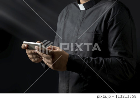 Young priest holding handphone, texting something isolated on black background. Young priest holding handphone, texting something isolated on black background. 113213559