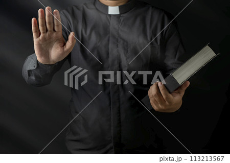 Young priest holding bible and raised hand giving forgiveness isolated on black background. 113213567