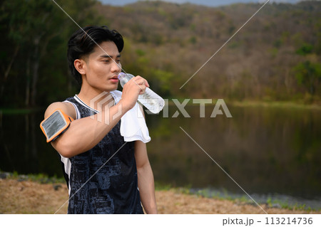 Athletic man standing near lake and drinking water after workout outdoors 113214736