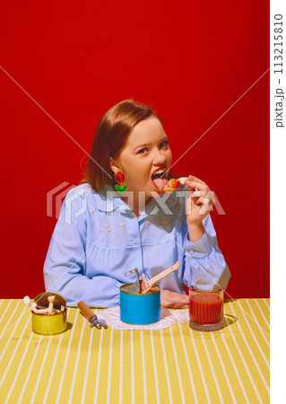 Young woman sitting at table with cans and eating doll with tomato juice against red background. Quirky dining 113215810