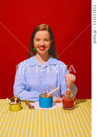 Smiling young woman sitting at table with cans and putting doll inside tomato juice against red background. Quirky dining Smiling young woman sitting at table with cans and putting doll inside tomato juice against red background. Quirky dining 113215811
