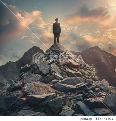 Silhouetted businessman stands on a rocky mountain peak against a majestic dawn sky, symbolizing success 113223103