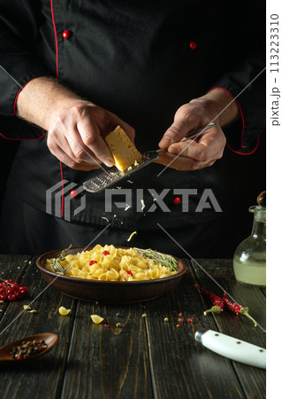A cook grates hard cheese into a plate of pasta with a hand grater. Working environment in restaurant kitchen with spices and kitchen utensils A cook grates hard cheese into a plate of pasta with a hand grater. Working environment in restaurant kitchen with spices and kitchen utensils 113223310