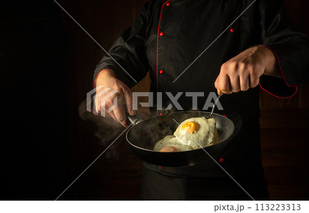 The cook prepares fried eggs in the saloon kitchen. A chef flips an egg in a steaming hot frying pan with a spatula. Low key concept of preparing delicious breakfast with space for recipe or menu The cook prepares fried eggs in the saloon kitchen. A chef flips an egg in a steaming hot frying pan with a spatula. Low key concept of preparing delicious breakfast with space for recipe or menu 113223313