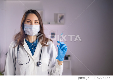 A young adult female doctor in a medical mask and white coat holds a syringe in her hands and looks at the camera 113224227
