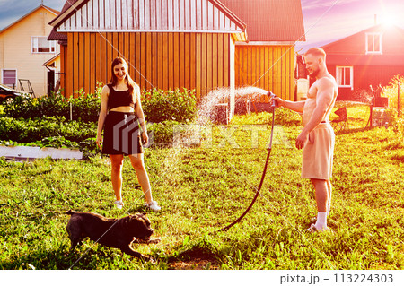 Man plays with dog, directing stream of water towards animal, young woman stands nearby and watches what is happening. Man plays with dog, directing stream of water towards animal, young woman stands nearby and watches what is happening. 113224303