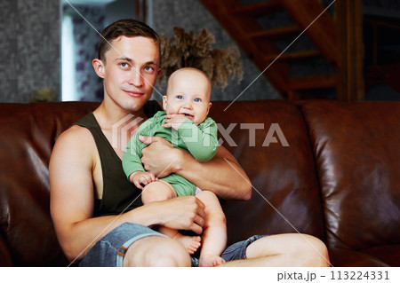 Caucasian man, 26 years old, holds his 6 month old baby daughter in his arms, sitting at home on leather sofa in living room. Caucasian man, 26 years old, holds his 6 month old baby daughter in his arms, sitting at home on leather sofa in living room. 113224331