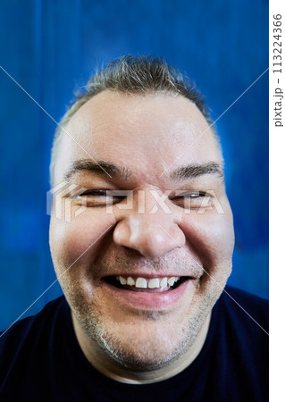 Full-mouth smile, close-up of face of large Caucasian man with wide smile on blue background, distorted by wide-angle lens, real photograph. Full-mouth smile, close-up of face of large Caucasian man with wide smile on blue background, distorted by wide-angle lens, real photograph. 113224366