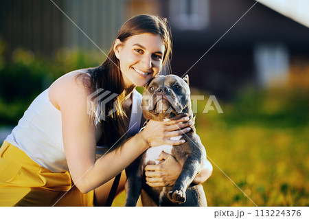 Young woman raised muzzle of American Bully to take joint portrait with her dog, white female is photographed with her pet on summer evening on lawn. Young woman raised muzzle of American Bully to take joint portrait with her dog, white female is photographed with her pet on summer evening on lawn. 113224376