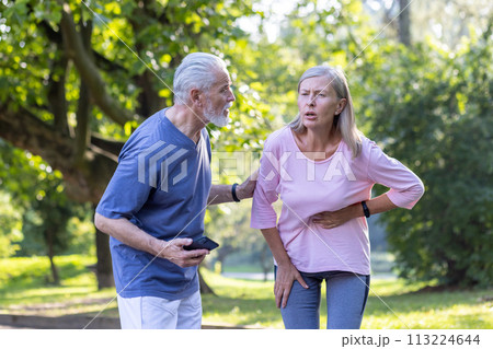 Senior couple doing sports and walking in the park, the woman feels severe pain in the side of the body and in the heart, the man tries to help, calls an ambulance on the phone. 113224644