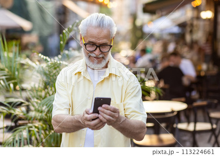 A happy senior gray-haired bearded man in a yellow shirt and glasses is walking in the city, and using a mobile phone with a smile. 113224661