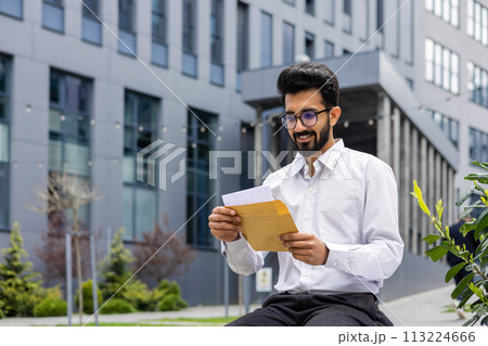 An Indian smiling young man in a white shirt sits outside a work building and reads a received letter and message in an envelope. An Indian smiling young man in a white shirt sits outside a work building and reads a received letter and message in an envelope. 113224666