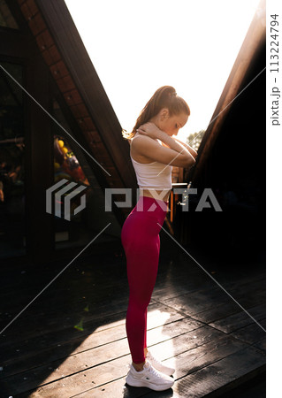 Vertical shot of sporty young woman in sportwear exercising and stretching neck muscles during warming up standing on background of country house on summer morning. Sporty female exercising outside. 113224794