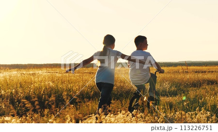 happy girl runs catching up with boy, child boy rides bicycle, son pedals, girl daughter plays airplane pilot, childhood dream airplane pilot fly, running sunset park, happy family kid, spinning wheel 113226751