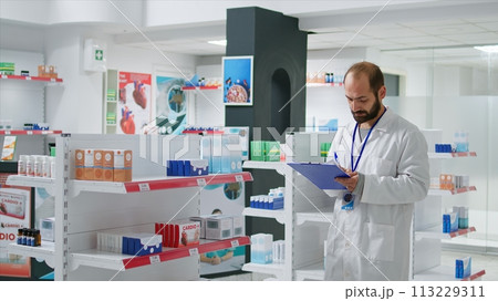 Pharmacy worker writing every medical product on inventory, creating long list with all pharmaceutics and medicaments in drugstore. Pharmacist ensuring variety of healthcare supplies. Camera 1. 113229311