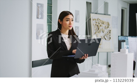 Design specialist reviewing measurements on 3d maquette model, working on remodeling property for a customer. Expert engineer looking at floor plans and architectural sketches on laptop. 113229339