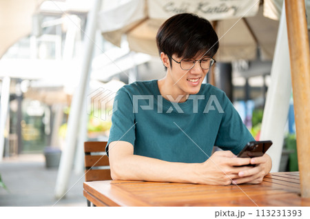 A smiling young Asian man wearing glasses sits at a wooden table outdoors, looking at his smartphone A smiling young Asian man wearing glasses sits at a wooden table outdoors, looking at his smartphone 113231393