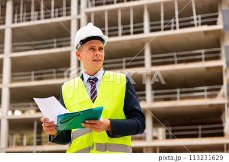 Architect in protective helmet and jacket with folder of documents on construction site 113231629
