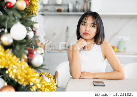 Sad young Asian girl waiting for phone call sitting at table by Xmas tree Sad young Asian girl waiting for phone call sitting at table by Xmas tree 113231829