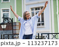 A woman standing in front of a building, arms outstretched. 113237173