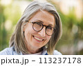 An older woman wearing glasses smiles directly at the camera. 113237178