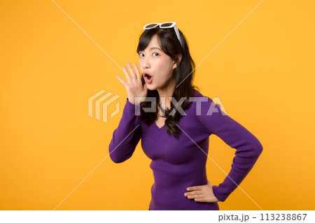 Portrait young beautiful asian woman 30s happy smile dressed in purple clothes shout to free space isolated on yellow studio background. Presenting product Beautiful girl act like telling to customer 113238867