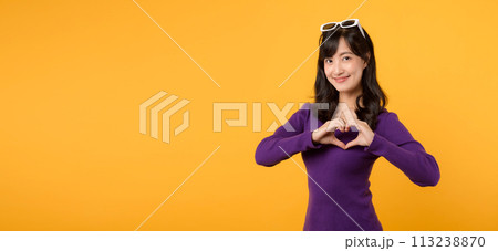Young cheerful asian woman 30s wearing purple sweater dress and sunglasses shapes heart gesture on yellow background. 113238870