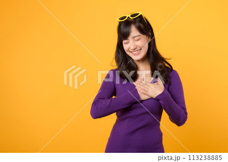 healthcare and wellness with an Asian young woman in her 30s, wearing a purple shirt. She showcases a hand gesture on her chest against a yellow background, embodying the concept of body wellness. 113238885