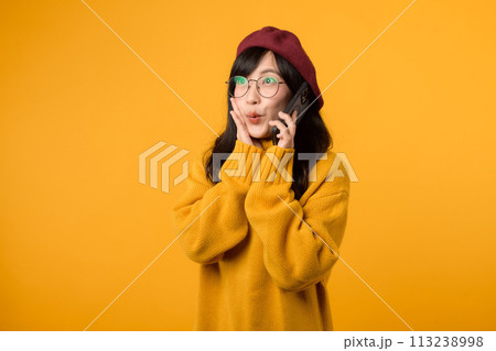 Female model in a trendy beret and yellow sweater, expressing joy while talking on her smartphone against a vibrant yellow background. Female model in a trendy beret and yellow sweater, expressing joy while talking on her smartphone against a vibrant yellow background. 113238998