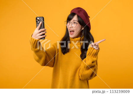 Portrait young beautiful 30s asian woman happy smile wearing yellow shirt and red hat pointing finger gesture to free copy space and using smartphone against yellow background. new application Portrait young beautiful 30s asian woman happy smile wearing yellow shirt and red hat pointing finger gesture to free copy space and using smartphone against yellow background. new application 113239009