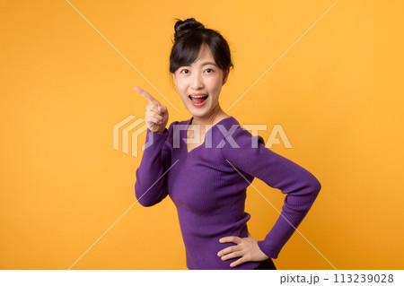 Portrait cheerful young asian woman 30s wearing purple shirt pointing finger to free copy space isolated on yellow background. Portrait cheerful young asian woman 30s wearing purple shirt pointing finger to free copy space isolated on yellow background. 113239028