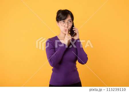 Mysterious Asian woman with purple shirt makes silence gesture presses index finger to lips shares secret with you holds mobile phone wears optical eyeglasses and warm jumper isolated over yellow wall 113239110