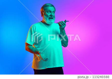 Emotional bearded senior man recording voice message via mobile phone on gradient blue-purple background in neon light 113240380